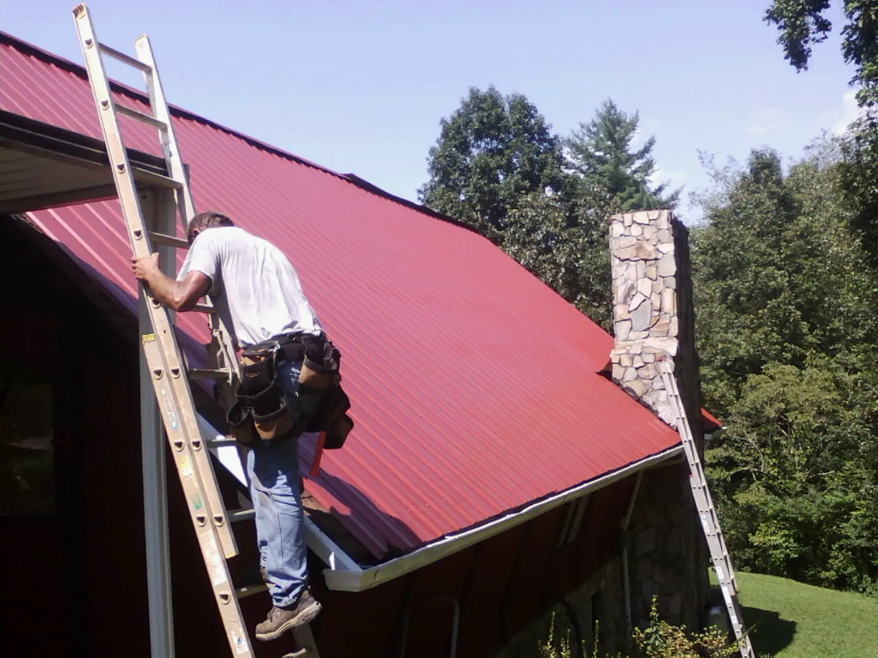 Roofer on a ladder beside a red metal roof and stone chimney — Soffit & Fascia Repair by Roofing Brothers in Carrolls